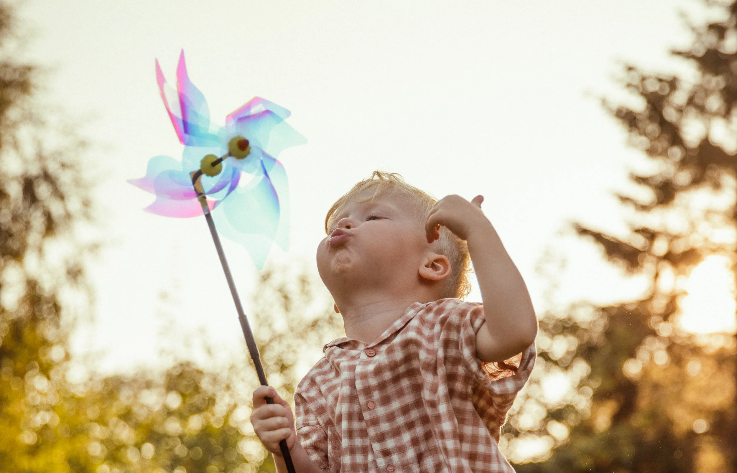 A young boy blowing on a colorful pinwheel outdoors during sunset.