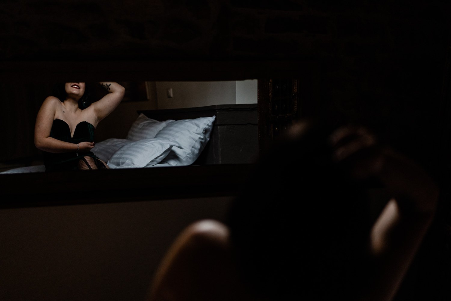 Une femme posant devant un miroir, reflétant une chambre sombre avec un lit.
