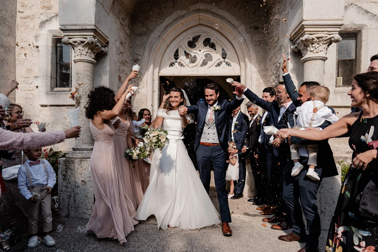 Un couple de mariés sortant de l'église, entouré d'invités qui les arrosent de bulles et de confettis. La mariée porte une robe blanche et tient un bouquet, le marié un costume bleu avec un nœud papillon. Les invités souriants célébrant l'instant dev