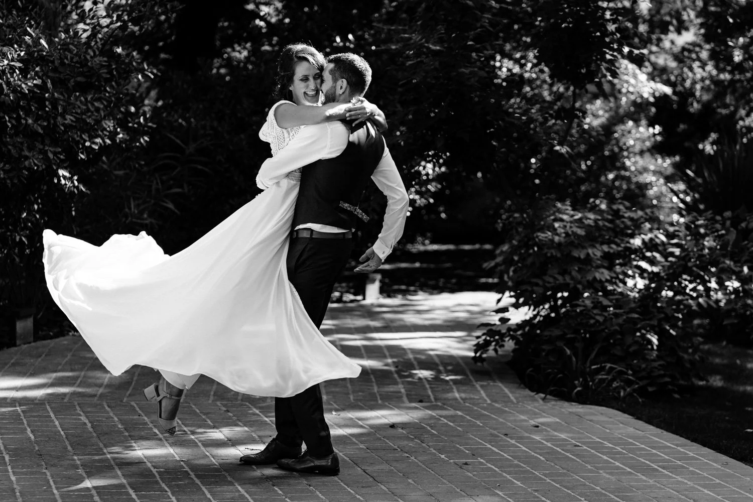 Un couple de mariés souriant et dansant en plein air, la femme en robe de mariage blanche, le homme en costume, entourés de verdure.