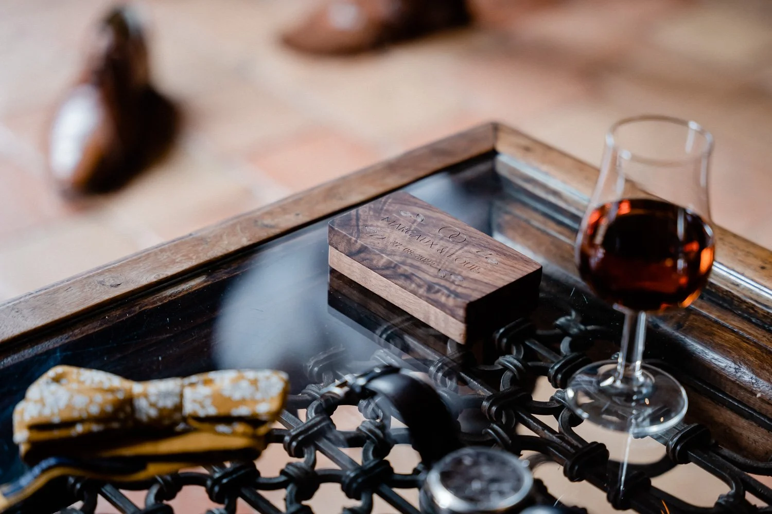 Une table en bois avec un verre de vin rouge, une boîte en bois gravée, une pochette en tissu jaune avec un nœud, et un plateau en metal.