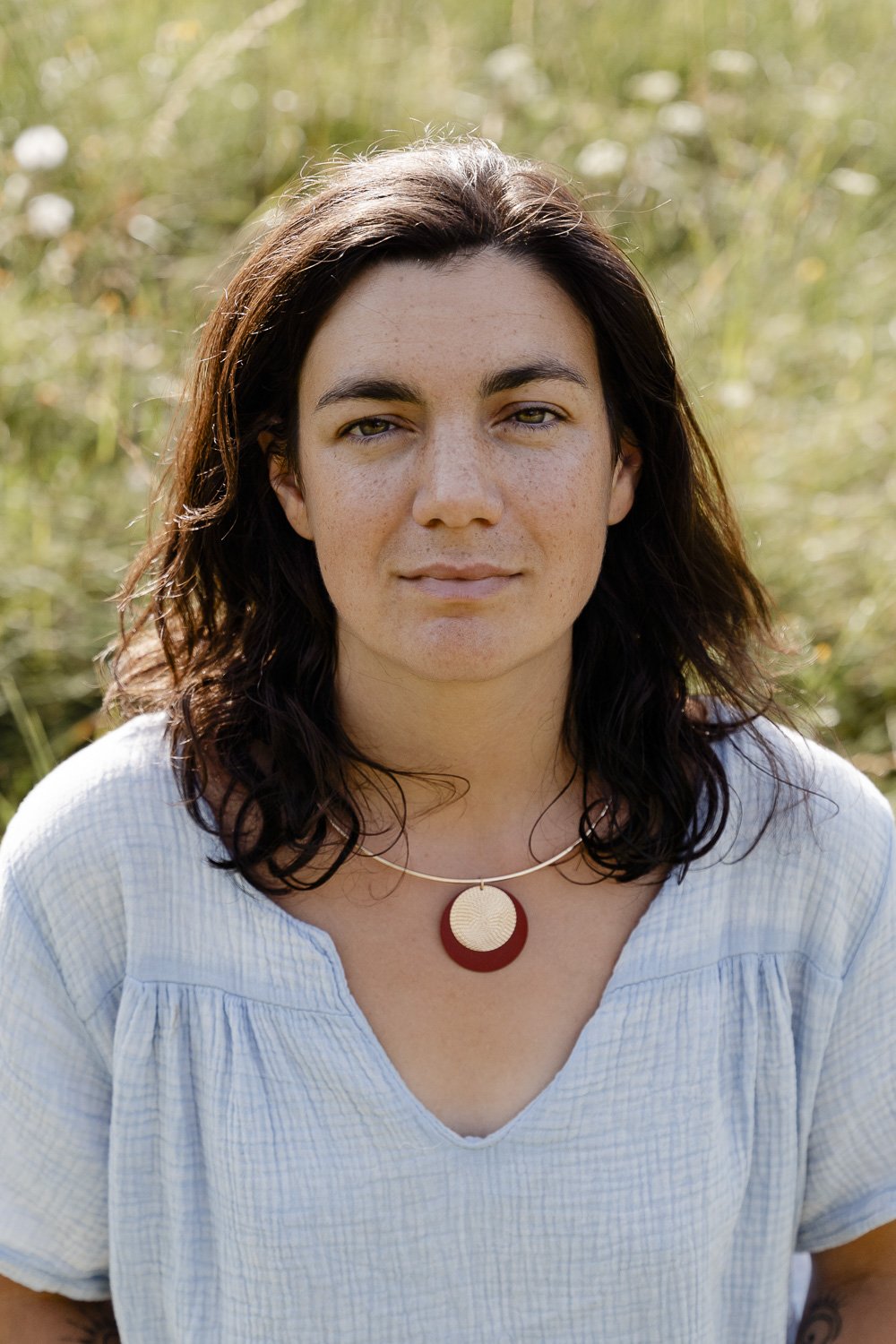 Une femme avec des cheveux bruns et un collier rouge, portant une blouse bleue, se tient dans un champ verdoyant.