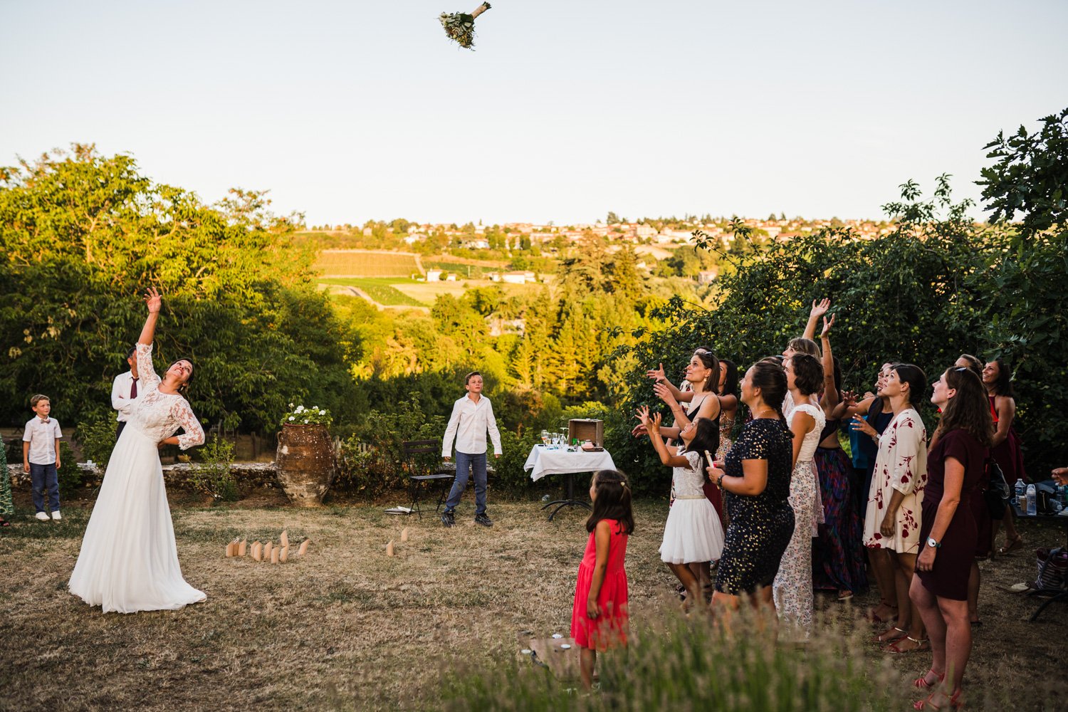 Une mariée lance un bouquet de fleurs en l'air, entourée de plusieurs personnes qui essaient de l'attraper, lors d'un mariage en plein air avec un paysage verdoyant en arrière-plan.