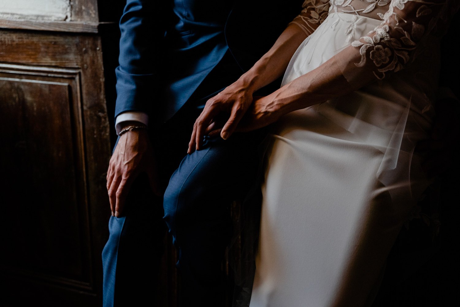 Partie inférieure de deux personnes assises, une en costume bleu et l'autre en robe blanche avec broderies en dentelle, leurs mains sont jointes sur leurs jambes, image sombre et intime.