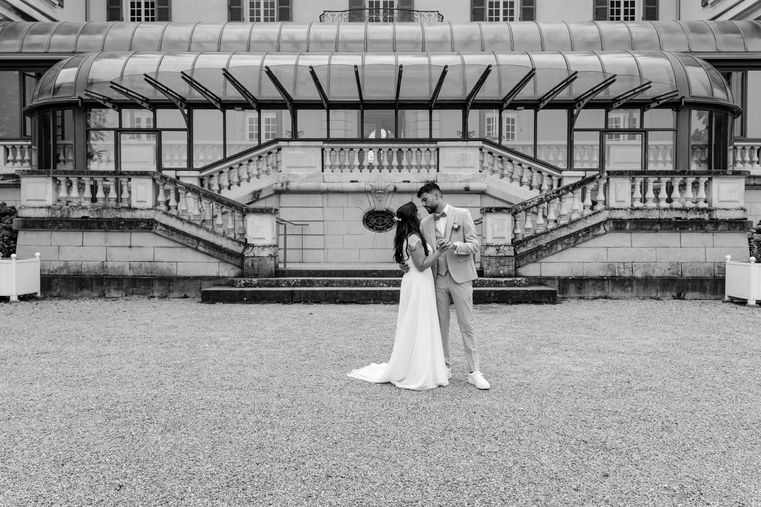 Un couple de mariés danse devant un bâtiment historique en pierre avec une véranda en verre, le tout en noir et blanc.