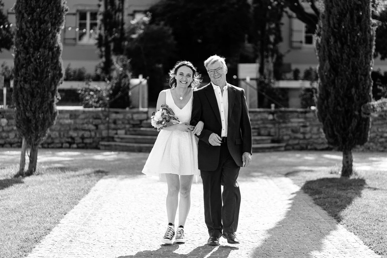 Une femme en robe de mariage tenait un bouquet, marchant avec un homme en costume lors d'une cérémonie en plein air.