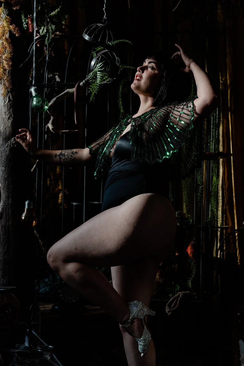 Une femme pose dans un décor sombre, portant une robe noire et un haut à paillettes vertes, avec des chaussures à talons brillantes. Elle a une coupe de cheveux courte et un tatouage sur le bras gauche.