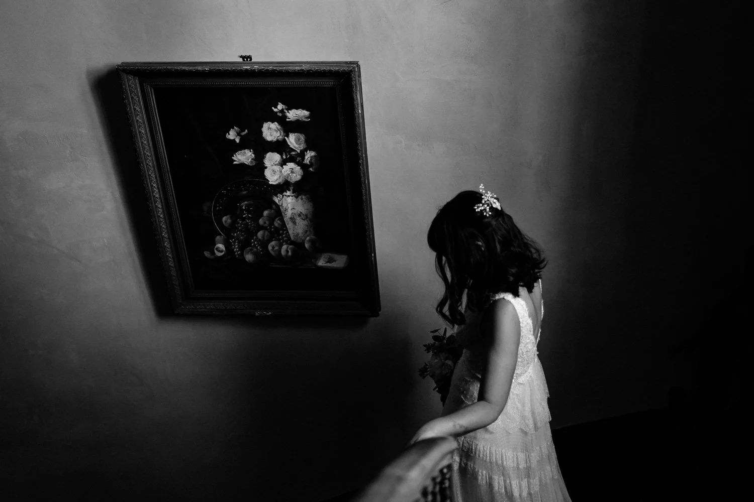 Une jeune fille en robe blanche, regardant un tableau accroché au mur, dans une pièce sombre en noir et blanc.