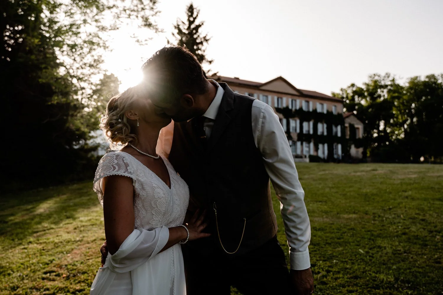 Un couple d'amoureux s'embrasse lors d'un mariage en extérieur avec un bâtiment en arrière-plan, en fin d'après-midi ou au coucher du soleil.