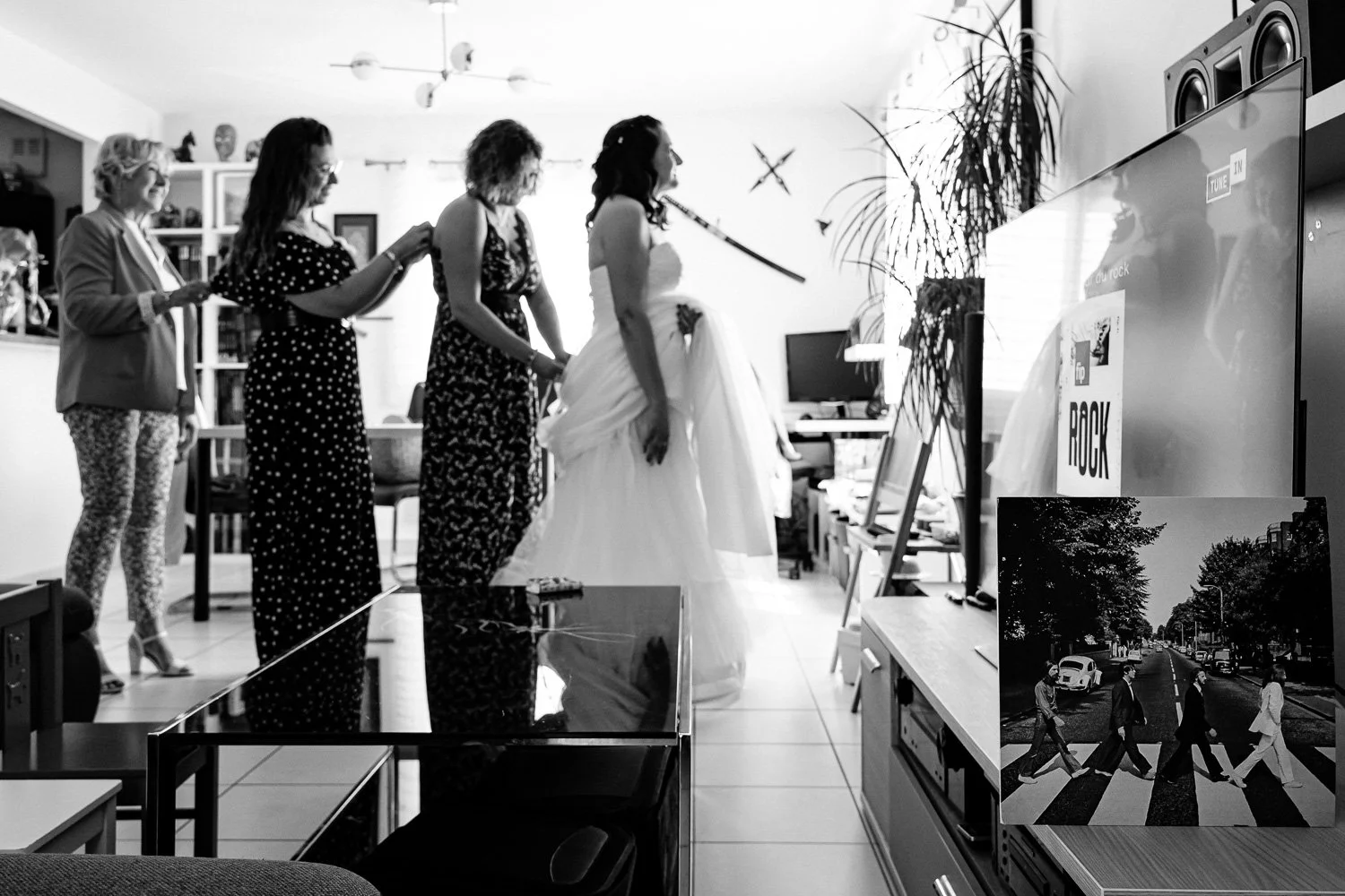 Une bride en robe blanche avec ses amies qui l'aident à se préparer, dans une pièce décorée avec un poster de la célèbre couverture de l'album 'Abbey Road' des Beatles. La scène est en noir et blanc.