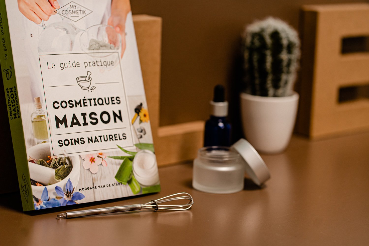 Livre "Cosmétiques Maison: Soins Naturels", flacon goutte-à-goutte, pot vide, fouet métallique, cactus en pot.