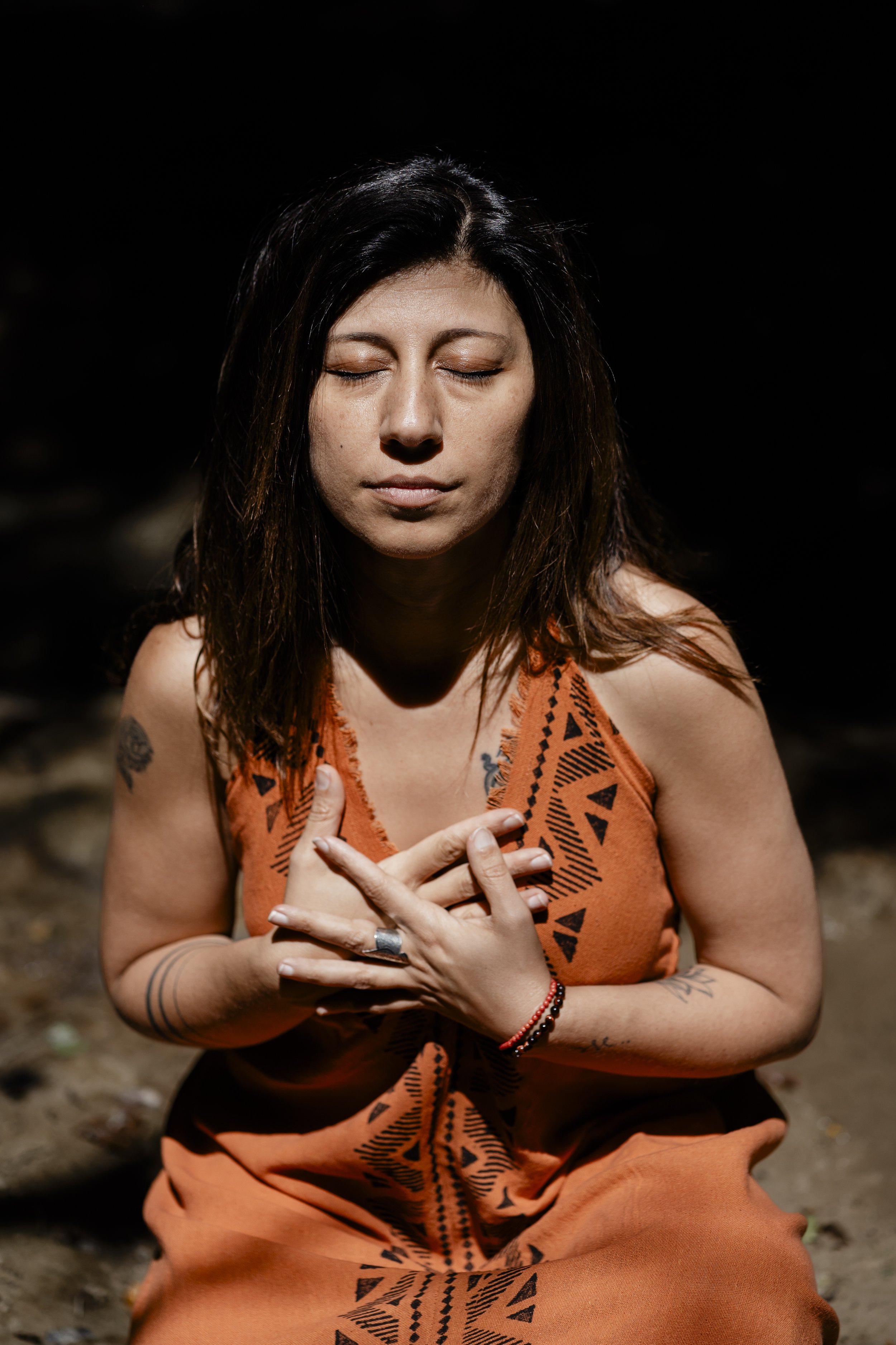 Femme méditant, yeux fermés, portant une robe orange avec des motifs tribaux, mains sur la poitrine.