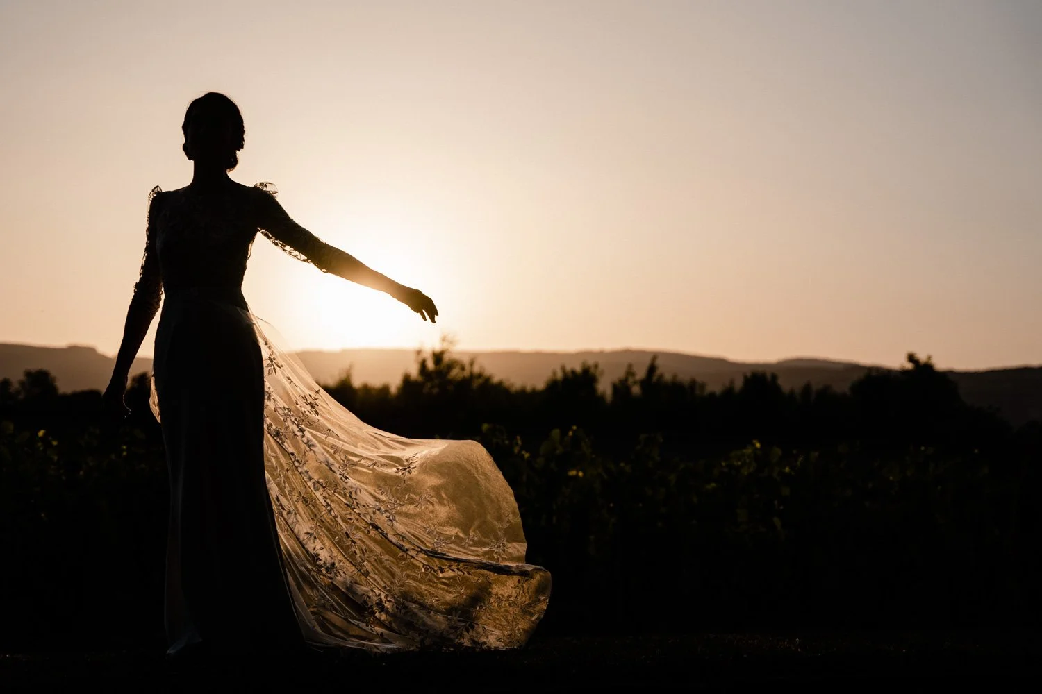 Silhouette d'une femme en robe longue et habillée de dentelle, marchant dans un champ au coucher du soleil.