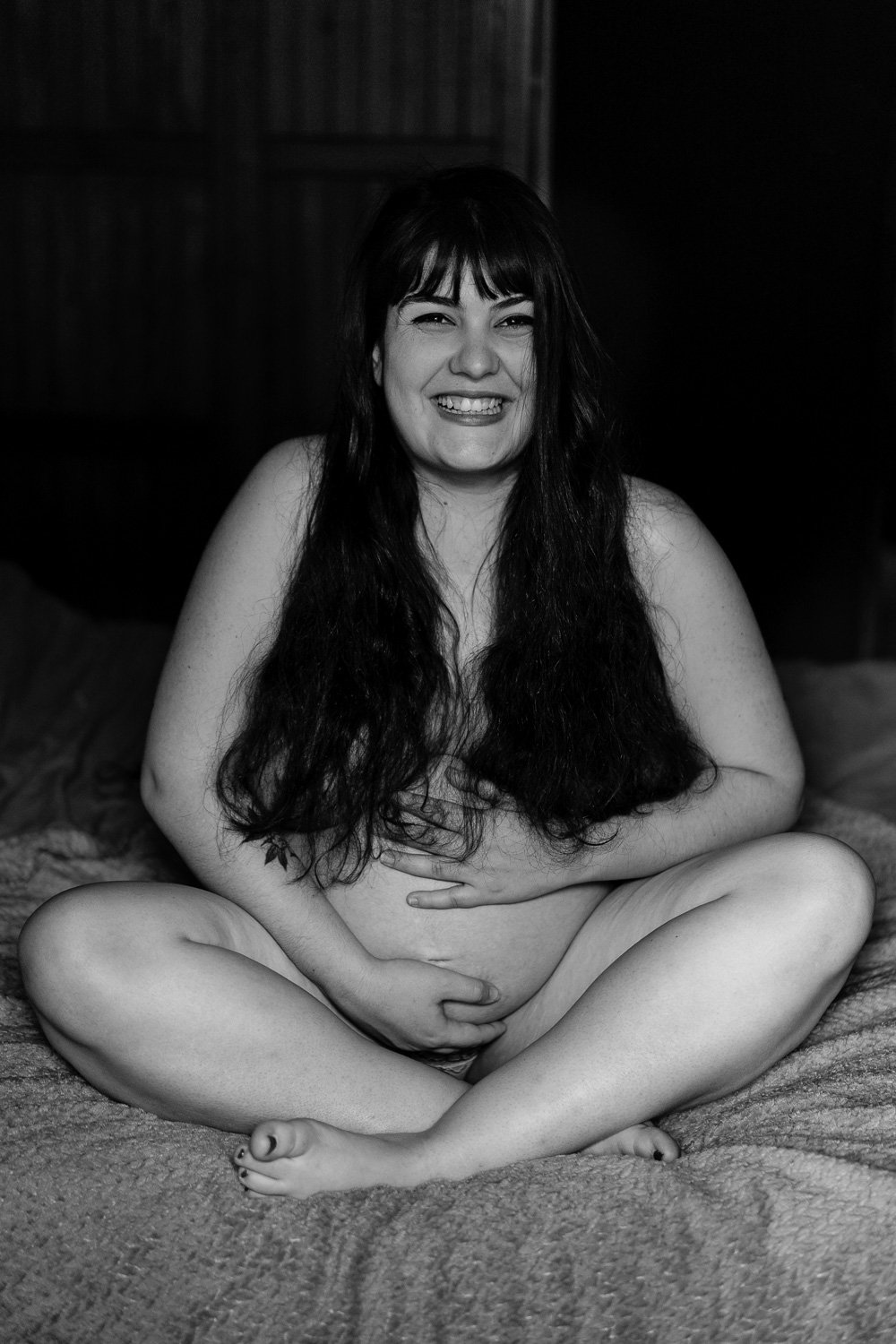 Femme enceinte souriante assise sur un lit, image en noir et blanc.