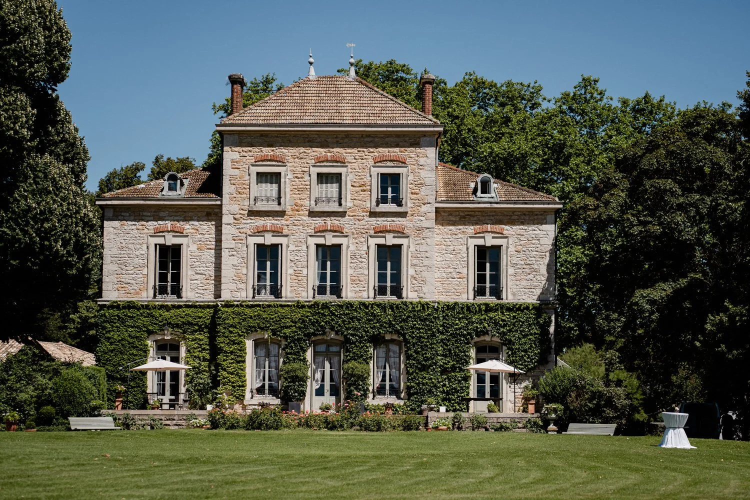 Maison en pierre de style traditionnel avec fenêtres à volets, entourée de verdure et de jardins bien entretenus, sous un ciel bleu clair.