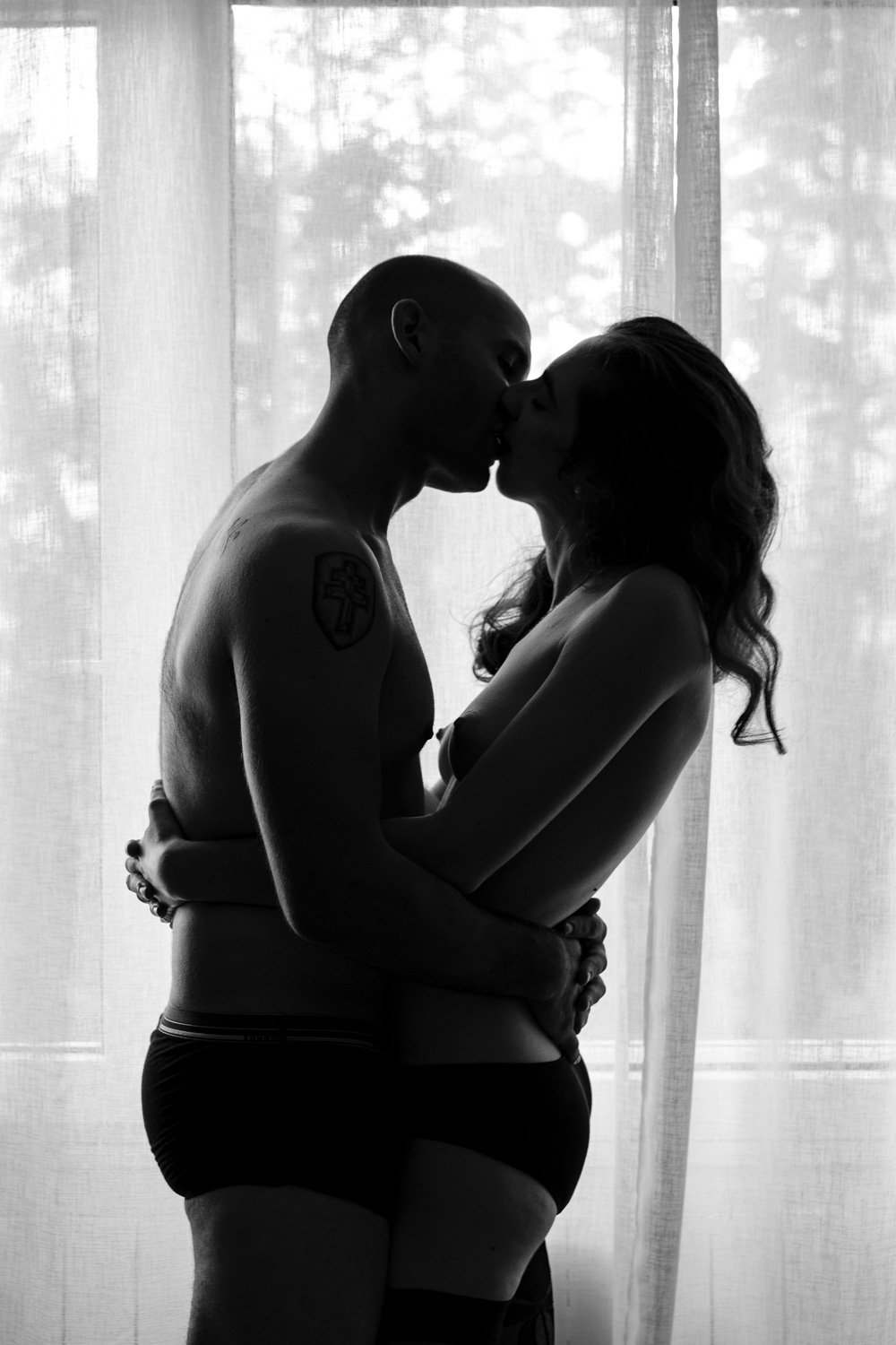 Un couple s'embrassant et s'enlaçant devant une fenêtre en silhouette, en noir et blanc.