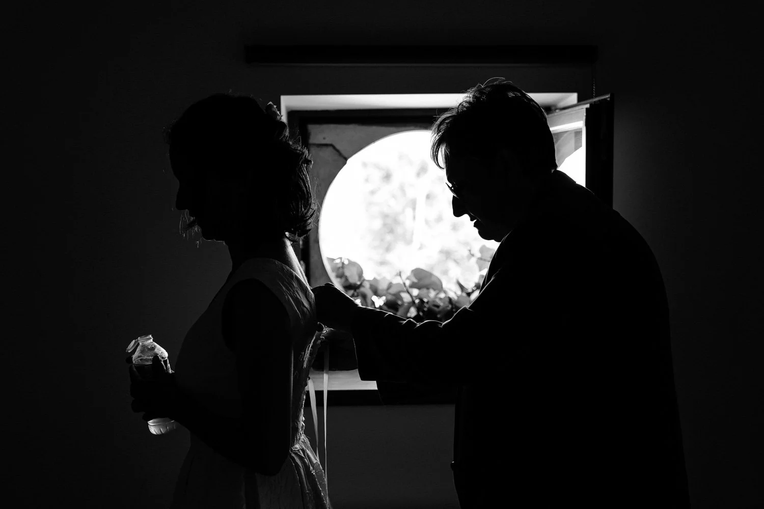 Silhouettes d'une femme et d'un homme dans une pièce sombre, avec une fenêtre en arrière-plan laissant entrer la lumière, capturant un moment intime.