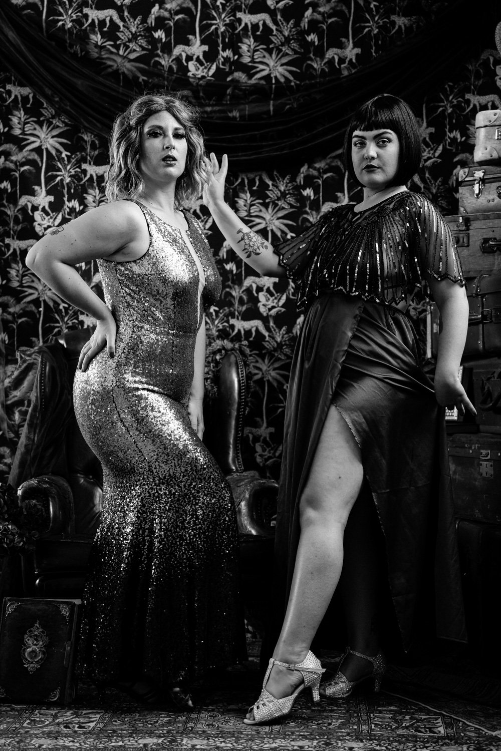 Deux femmes posant dans une pièce au décor vintage, portant des robes de soirée élégantes, l'une en robe à paillettes et l'autre en robe à perles. Le fond est orné de motifs floraux et d'anciennetés.