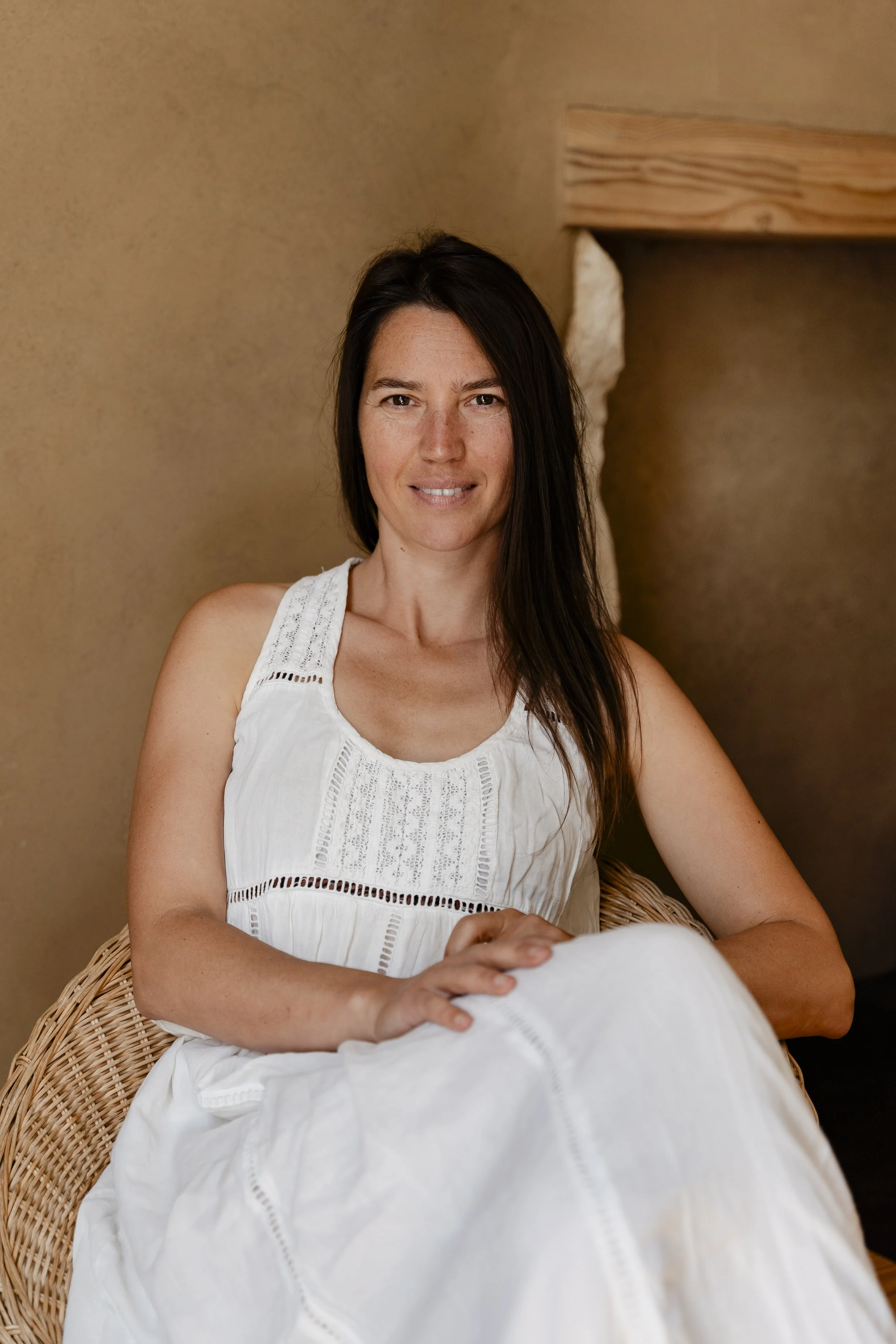 Une femme aux cheveux bruns et longs, portant une robe blanche en crochet, assise dans un fauteuil tressé, à l'intérieur d'une pièce avec un mur beige et une cheminée en pierre en arrière-plan.