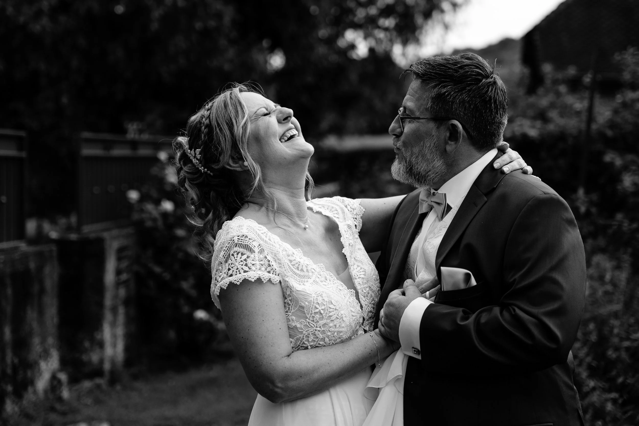 Un couple de mariage en noir et blanc, la femme rit joyeusement, et l'homme, avec des lunettes, la regarde affectueusement, dans un décor extérieur.