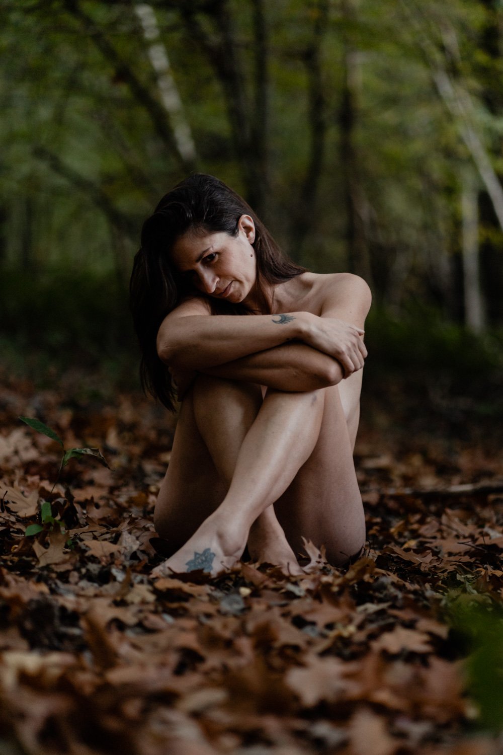 Femme assise dans une forêt, entourée de feuilles mortes, les bras croisés sur ses genoux.