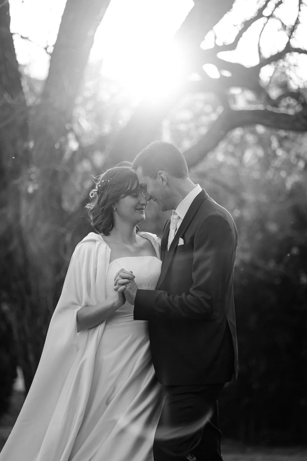 Un couple en mariage, ils se tiennent par la main, face à face, dans un cadre naturel avec des arbres, en noir et blanc, avec le soleil en arrière-plan.