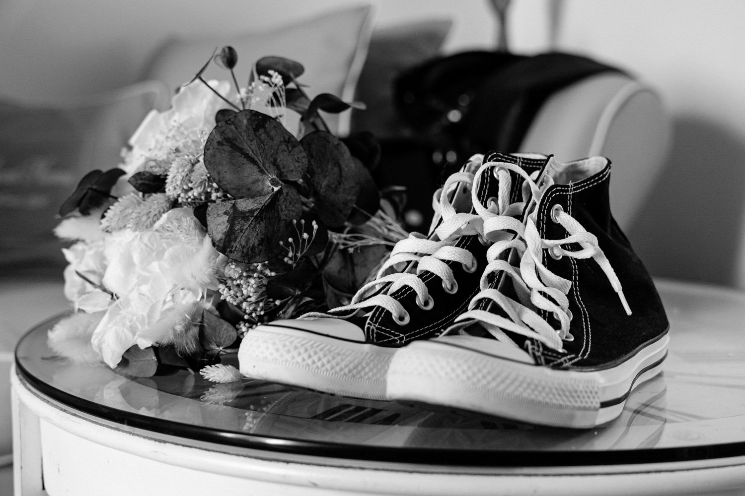Une paire de baskets noires avec des lacets blancs posée sur une table en bois, à côté d'un bouquet de fleurs, dans une pièce intérieure en noir et blanc.