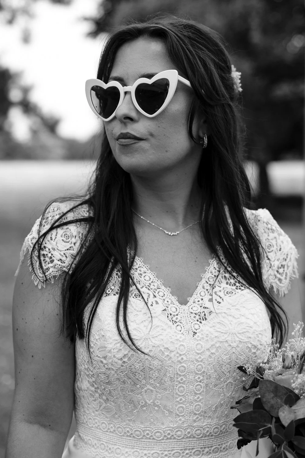Une femme portant des lunettes de soleil en forme de cœur, avec une robe en dentelle, tenant un bouquet, en extérieur, en noir et blanc.