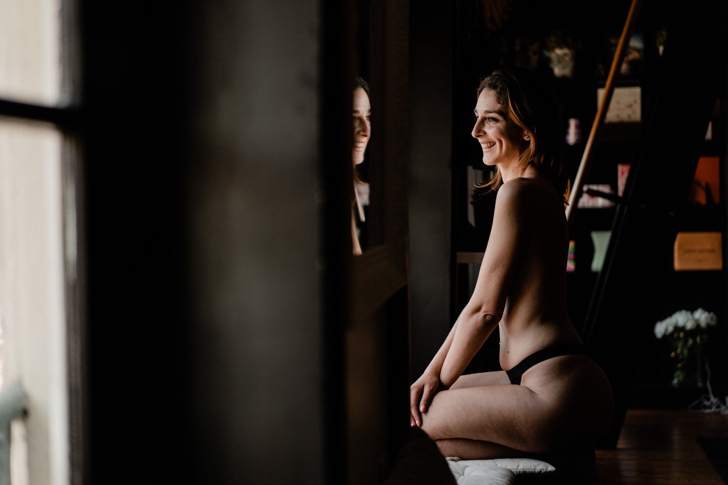 Une femme en sous-vêtements noirs est assise près d'une fenêtre, avec un sourire, illuminée par la lumière naturelle.