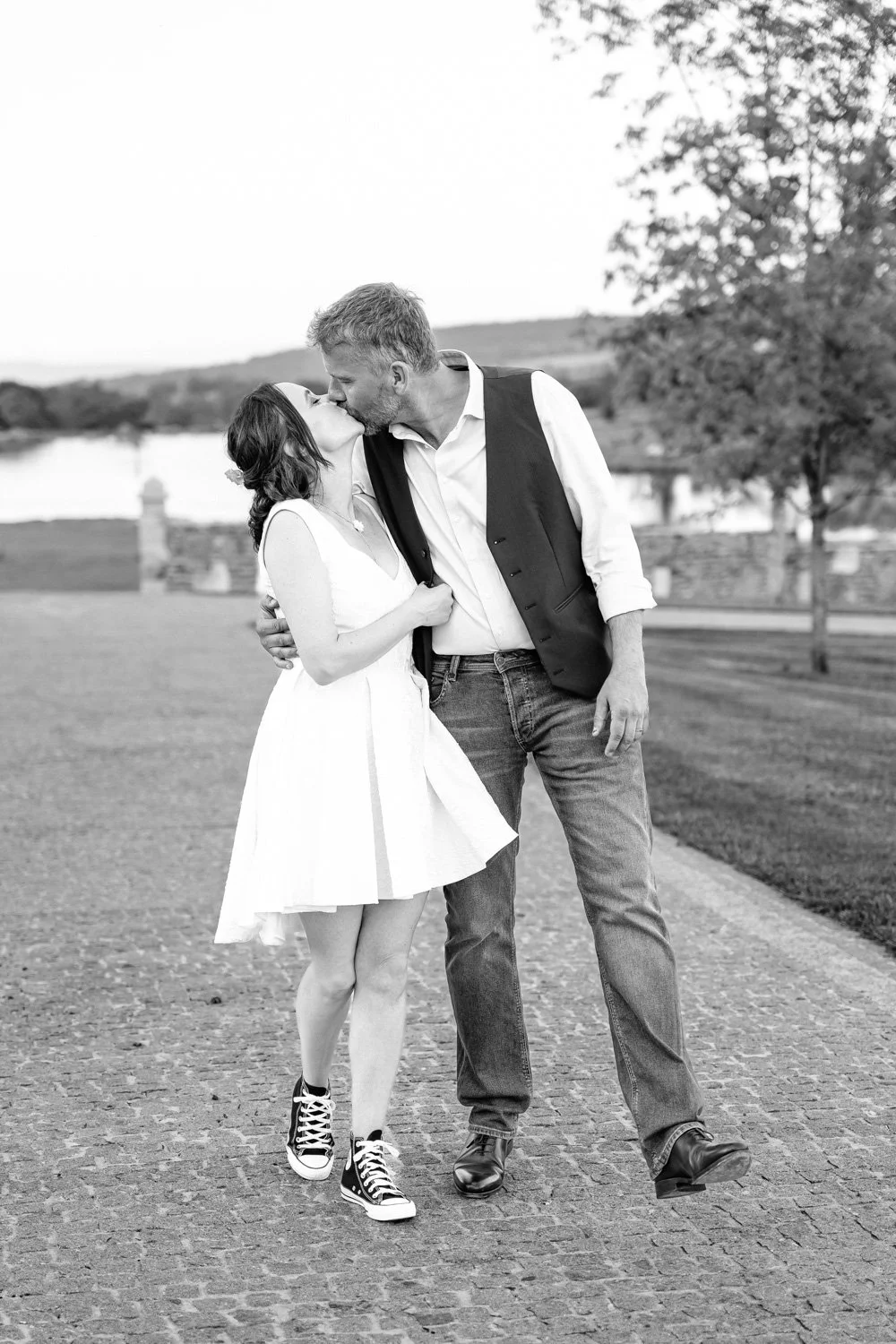 Un couple s'embrasse et échange un baiser dans un parc au bord de l'eau, en noir et blanc.
