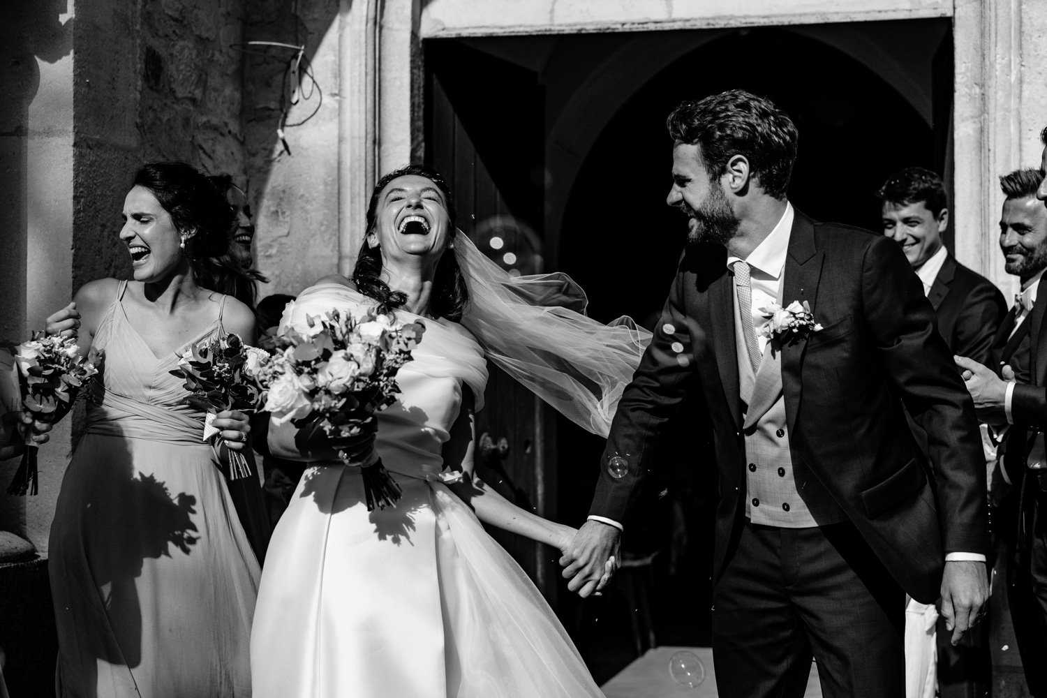Un couple de mariés heureux sort de l'église, tenant des bouquets de fleurs, entouré d'amis souriants.