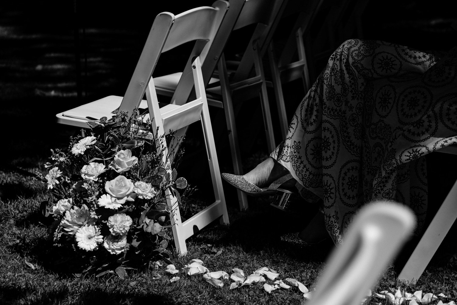 Chaise pliante à côté d'un bouquet de fleurs sur le sol en plein air, une personne assise portant une robe à motifs et des chaussures à talons. Photographie en noir et blanc.