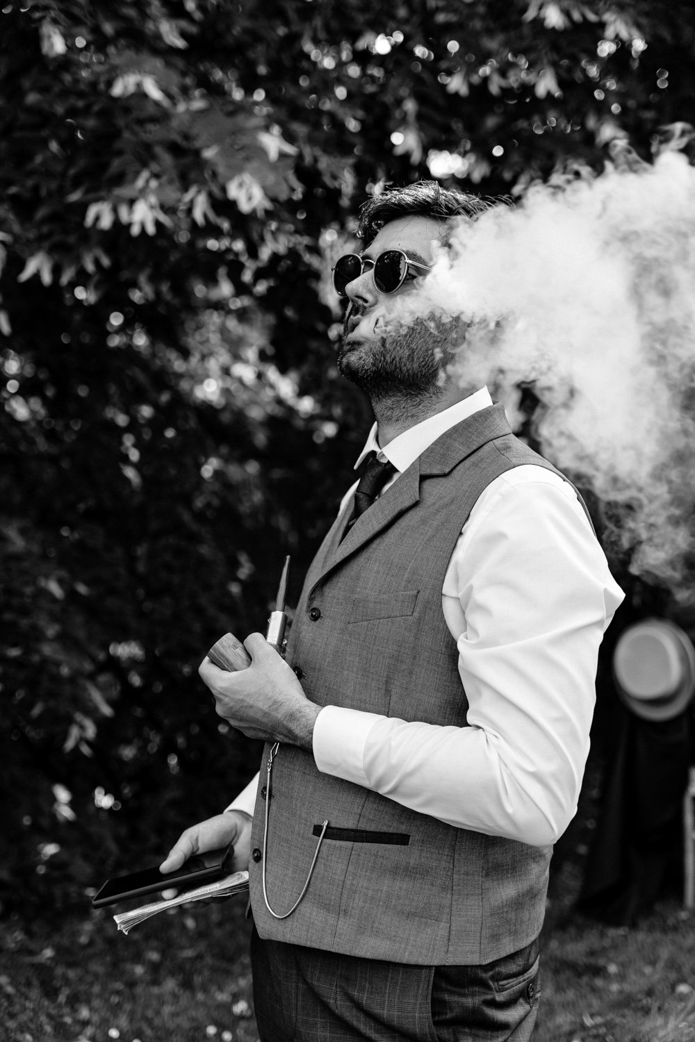 Homme en costume fumant avec une pipe, lunettes de soleil, tenant un livre et un téléphone, arborant une chaîne de montre, fond de feuillage.