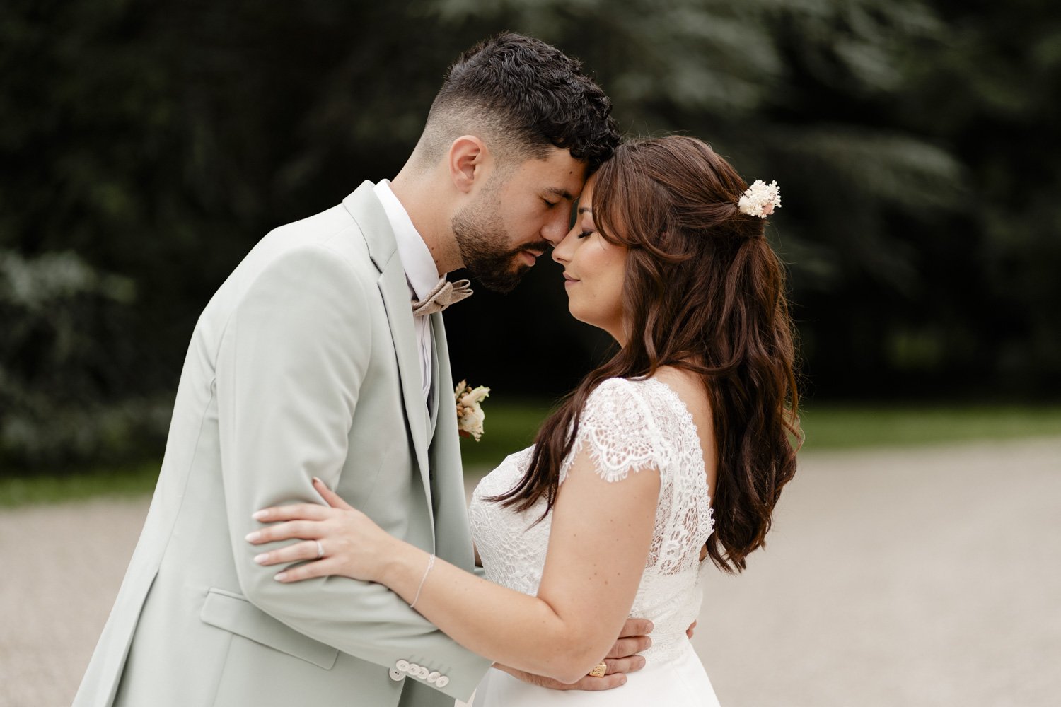 Un couple habillé en tenue de mariage s'enlace tendrement, l'homme en costume clair et la femme en robe de mariée avec dentelle, dans un cadre extérieur verdoyant.