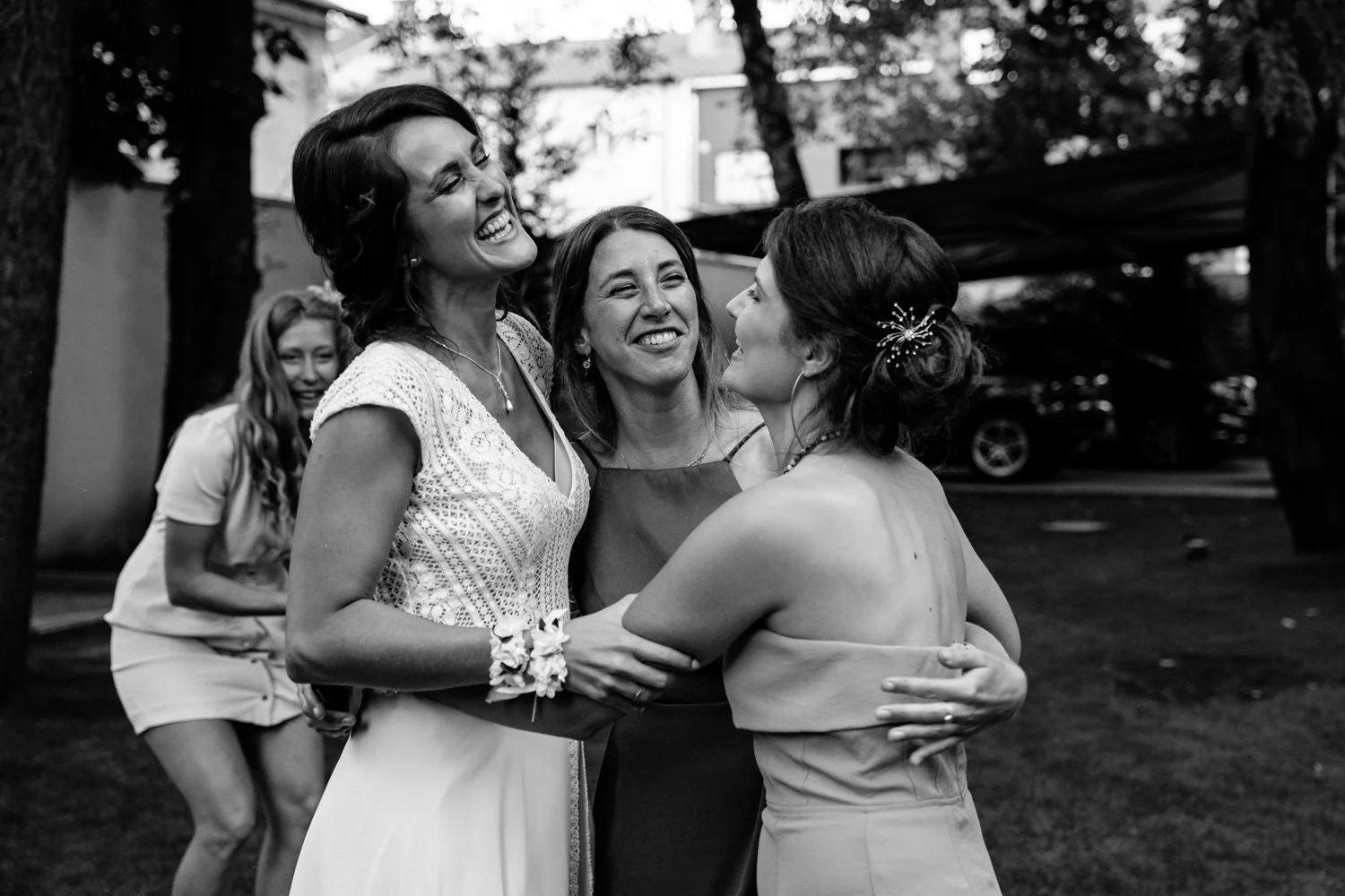 Groupe de quatre femmes souriantes, deux en bras croisés, en plein partage d'un moment joyeux en plein air, avec arbres et voitures en arrière-plan.