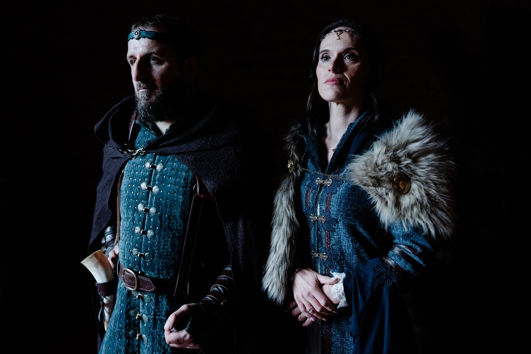 Deux personnes en costumes médiévaux, un homme et une femme, avec des vêtements ornés et un éclairage dramatique.