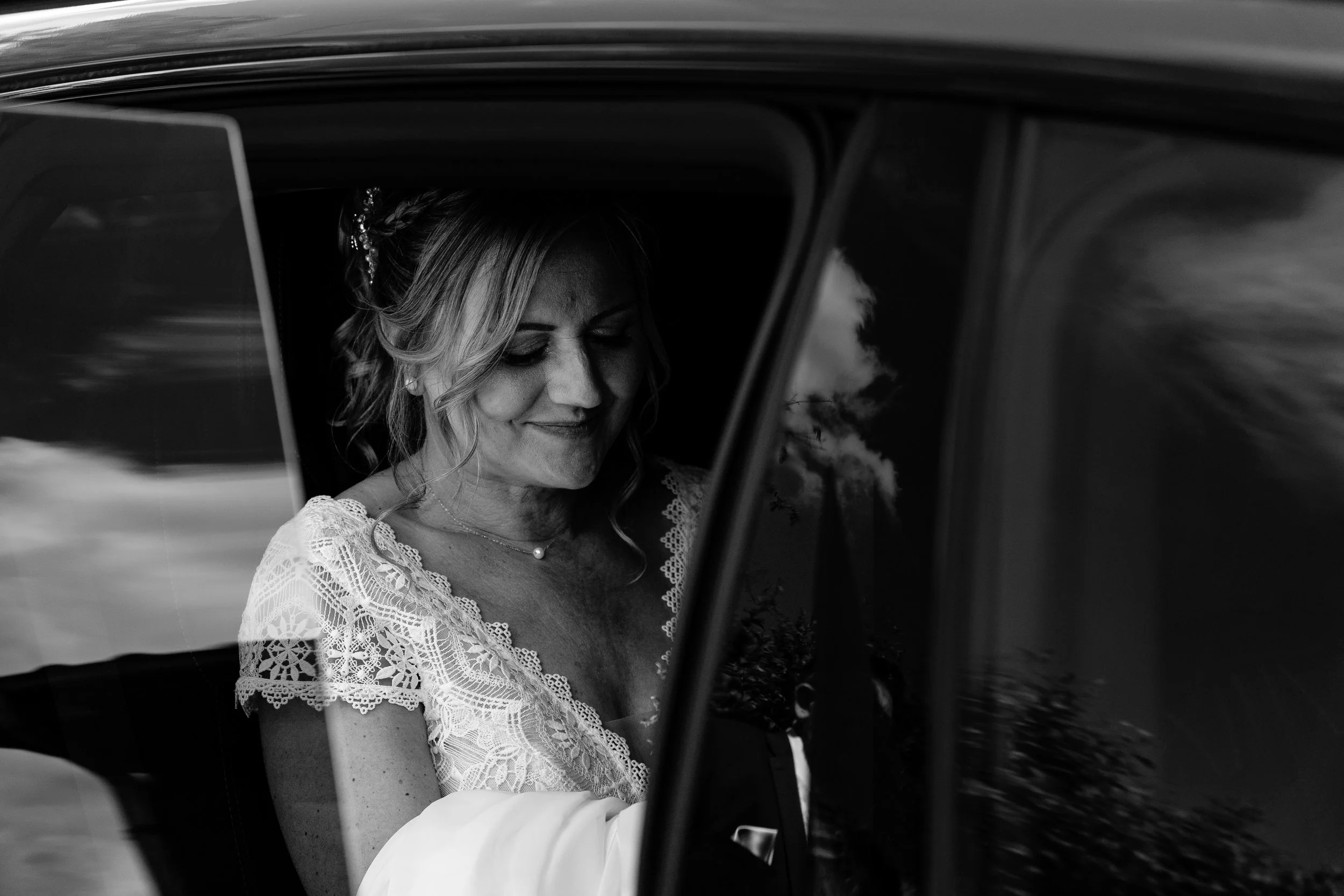 Une femme souriante dans une robe de mariée, vue à travers la fenêtre d'une voiture, en noir et blanc.