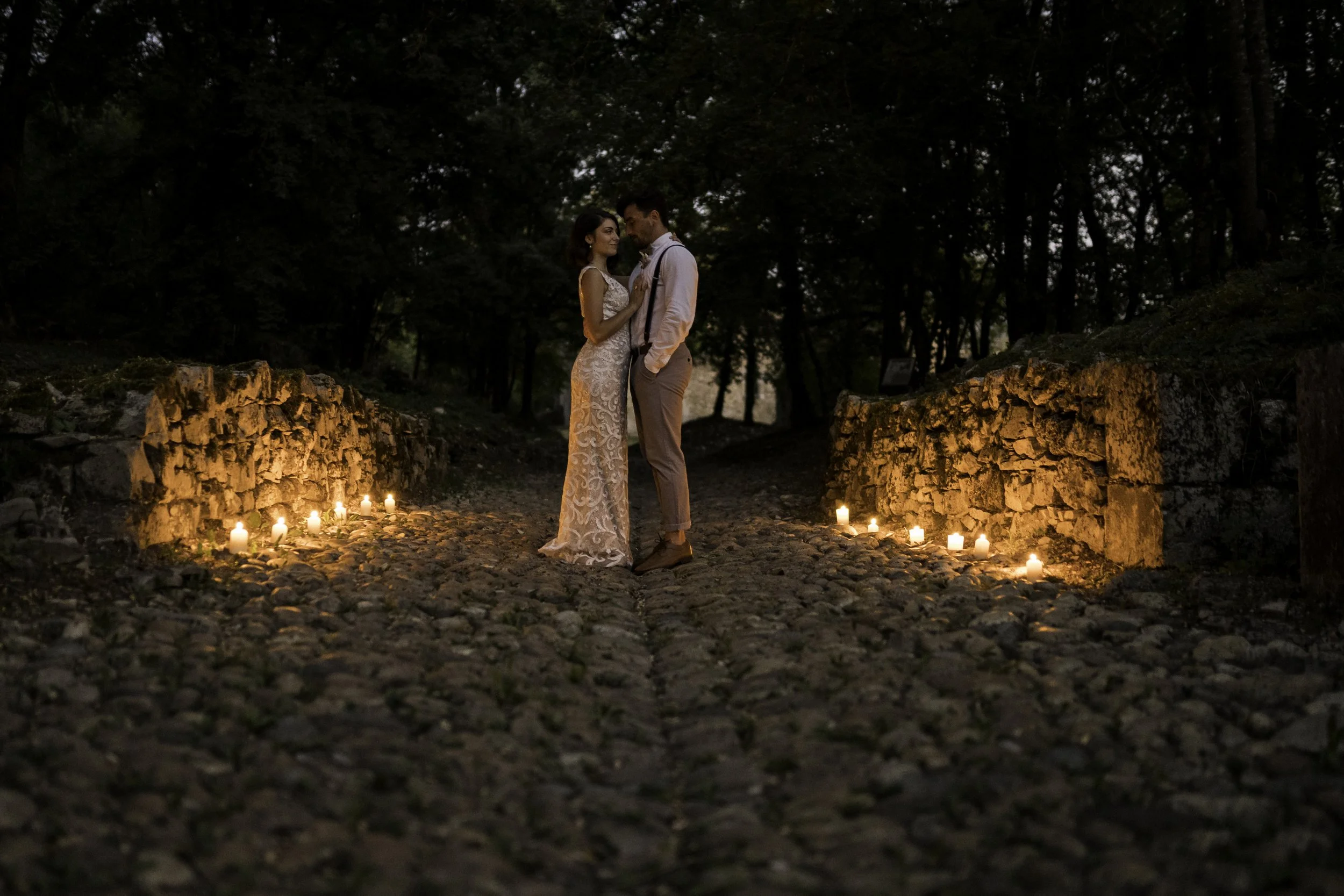 Un couple en robe de mariage et costume se tient sous un ciel crépusculaire, entouré de bougies allumées sur un chemin pavé dans une forêt, créant une ambiance romantique.