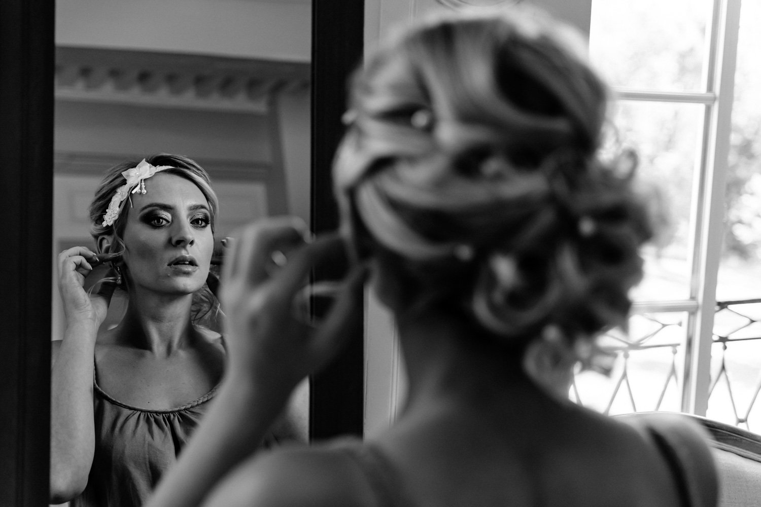 Une femme regarde son reflet dans un miroir en se maquillant, avec un décor intérieur en arrière-plan et une fenêtre à l'extérieur.