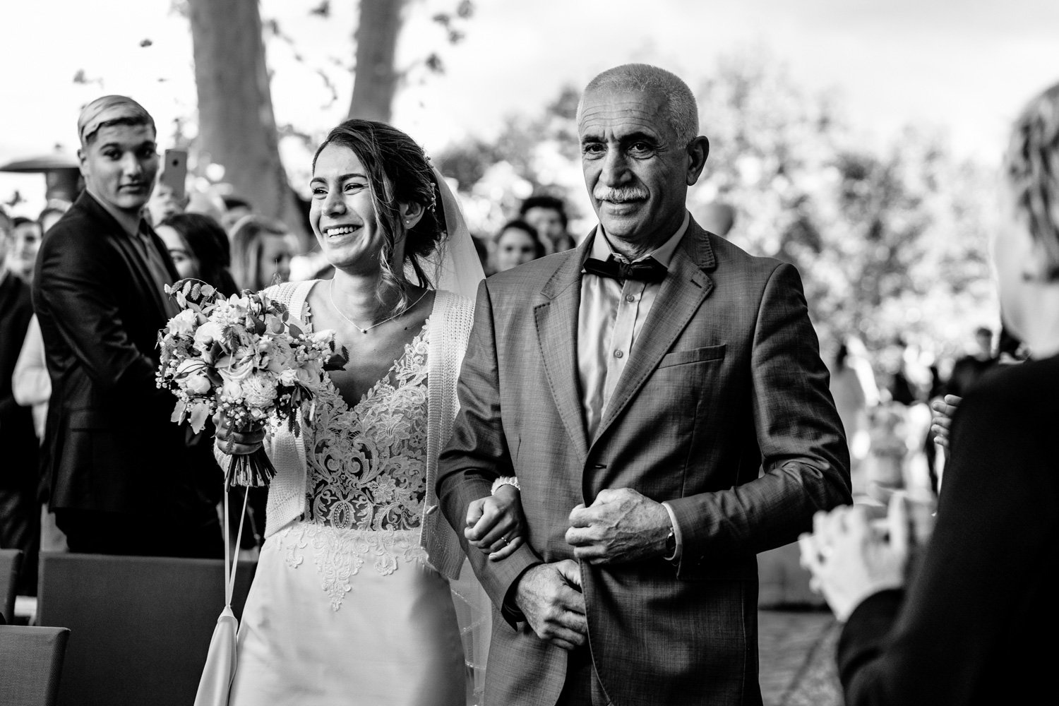 Mariée souriante avec un bouquet marchant au bras d'un homme âgé, probablement pendant une cérémonie de mariage.