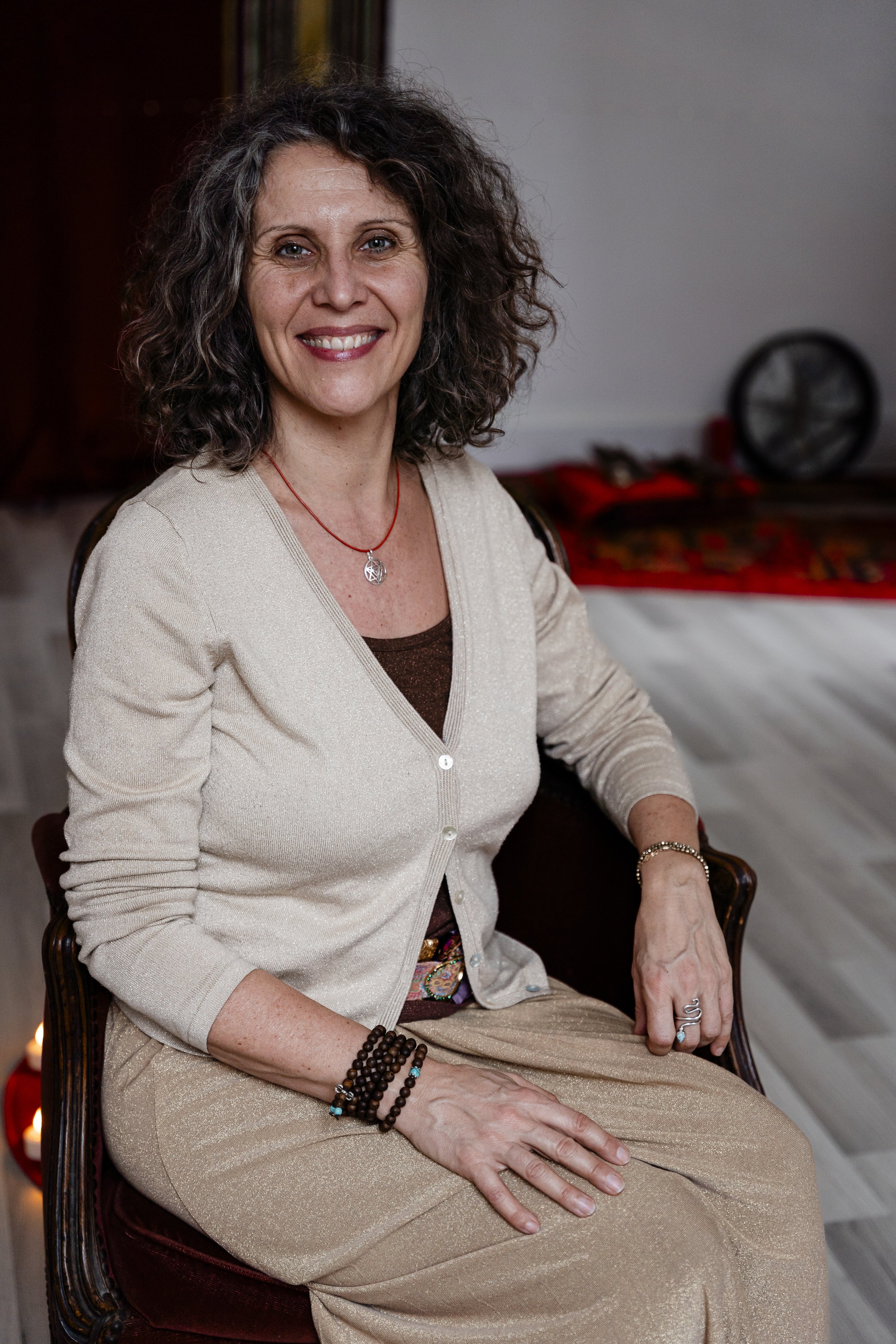 Femme souriante assise sur une chaise, portant un cardigan beige et un collier, avec un bracelet au poignet.