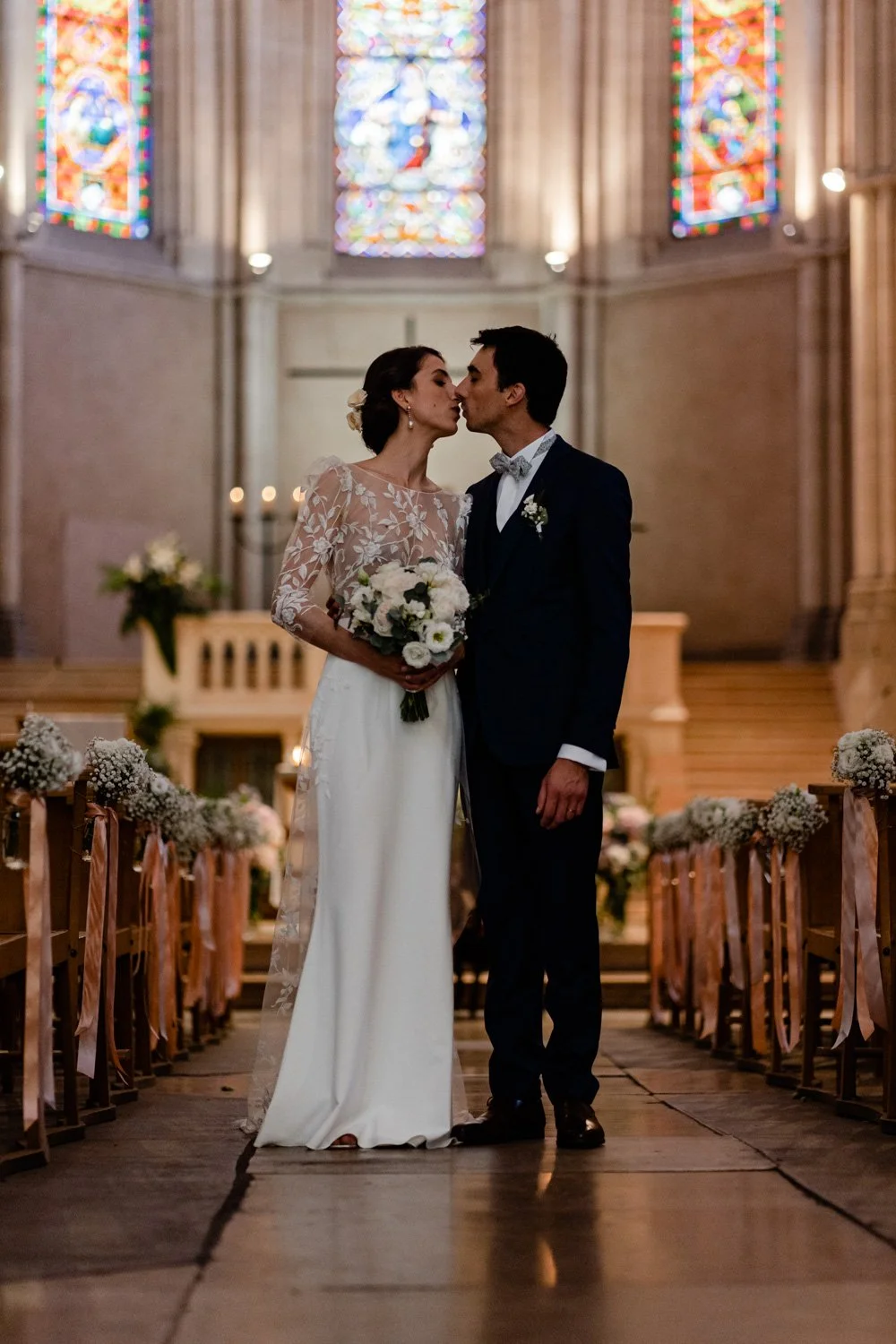 Un couple de mariés dans une église, s'apprêtant à s'embrasser, avec des vitraux colorés en arrière-plan, décorée de fleurs et de rubans pour leur mariage.