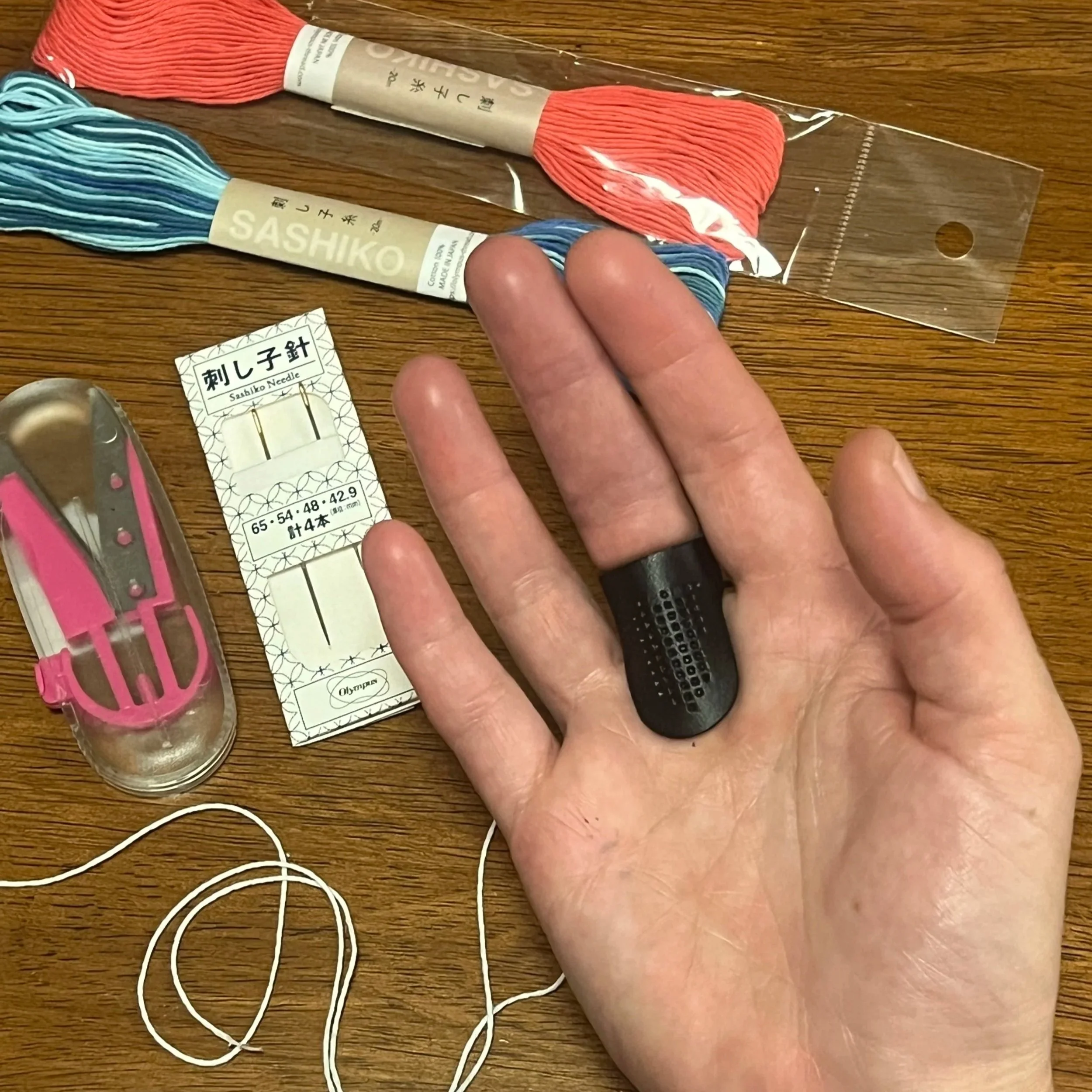 Hand wearing sashiko thimble with threads, scissors, and needles on a table.