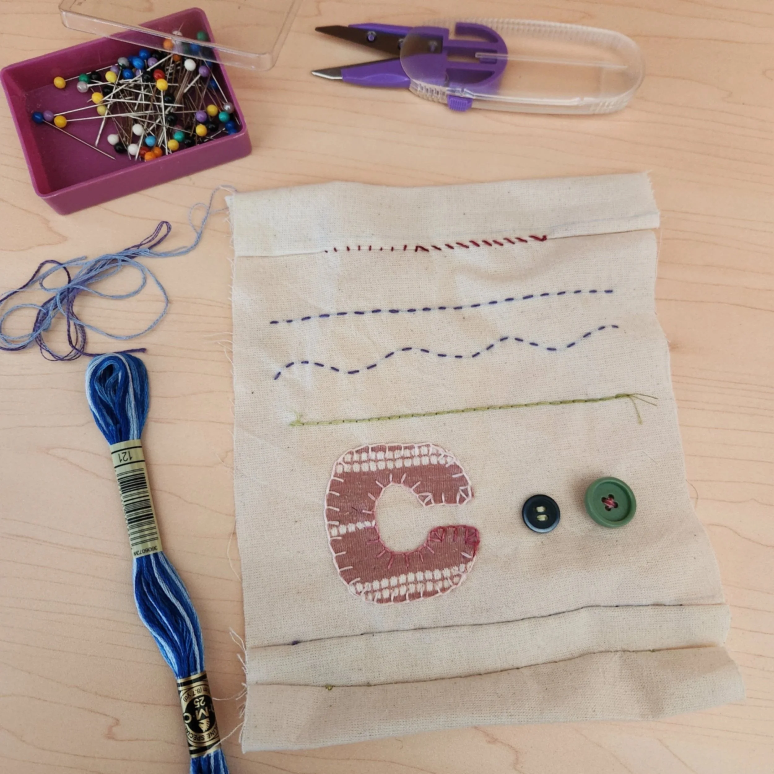 Fabric sampler showing stitched rows, hand-sewn buttons, and a large appliquéd letter C, surrounded by thread, pins, and scissors.