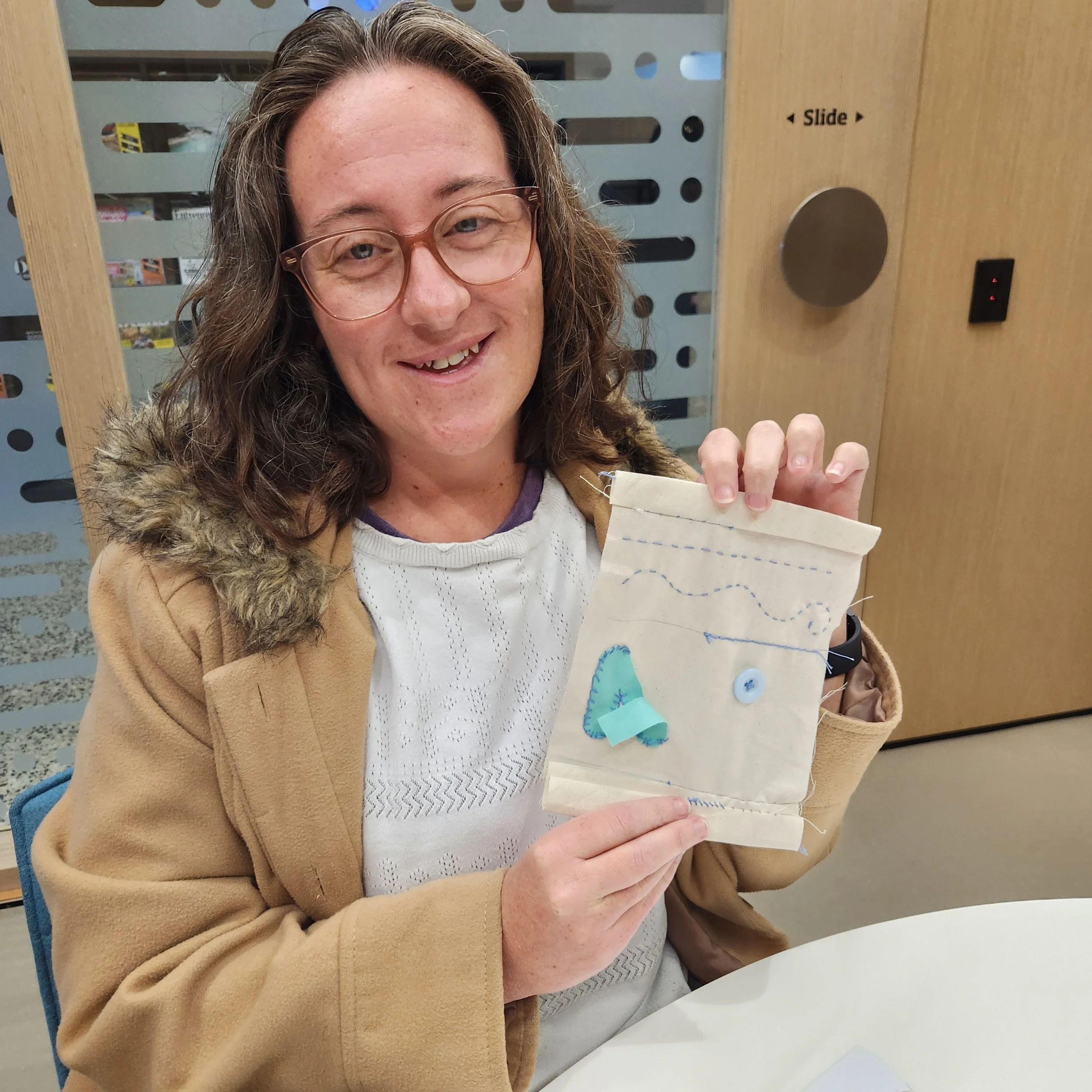 Smiling workshop participant holding a calico sampler with blue stitches, a sewn button, and an appliquéd fabric shape.