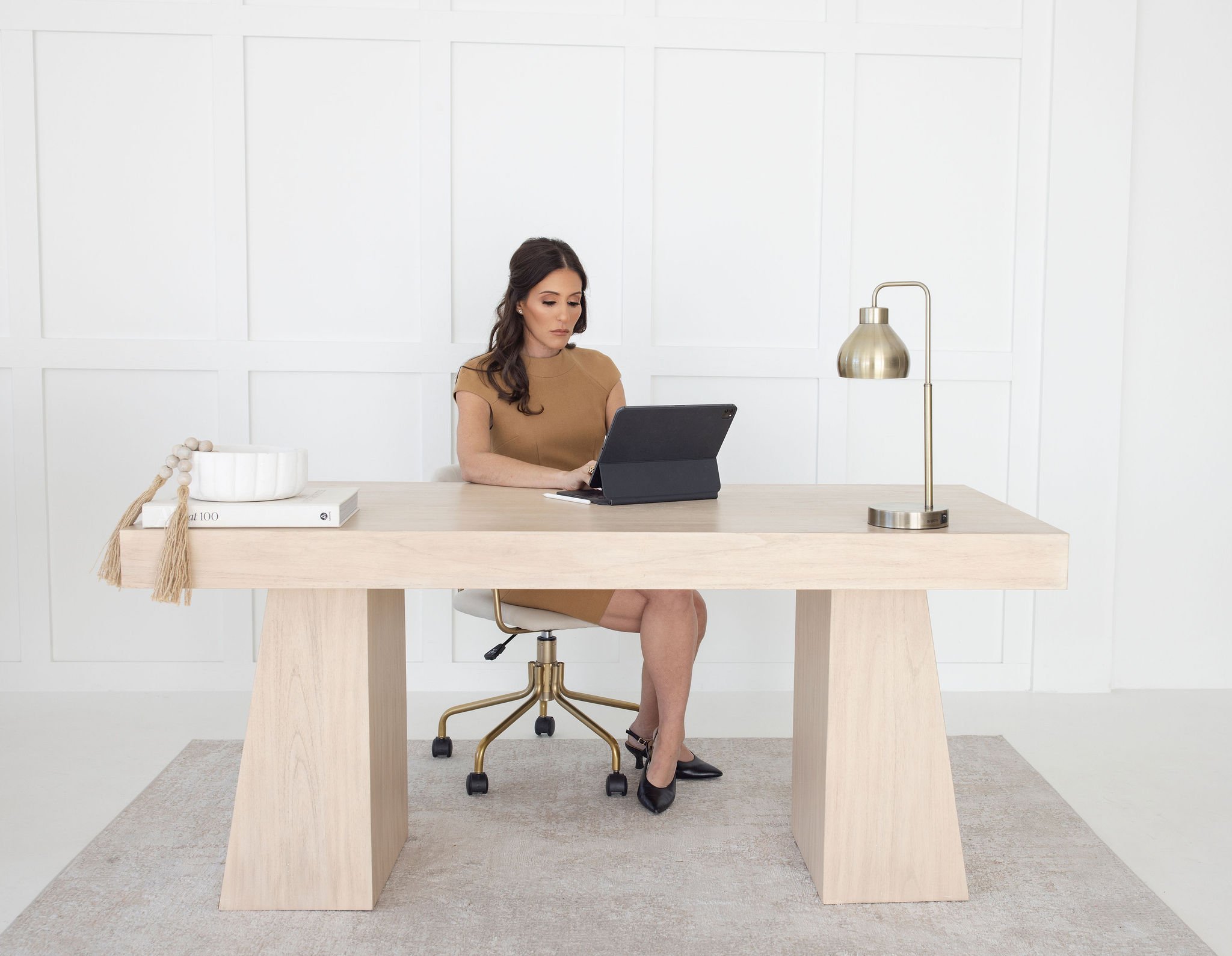 A woman sitting at a light wooden desk in a white room, working on a laptop, with a gold desk lamp, a stack of books, and a decorative bowl on the desk.