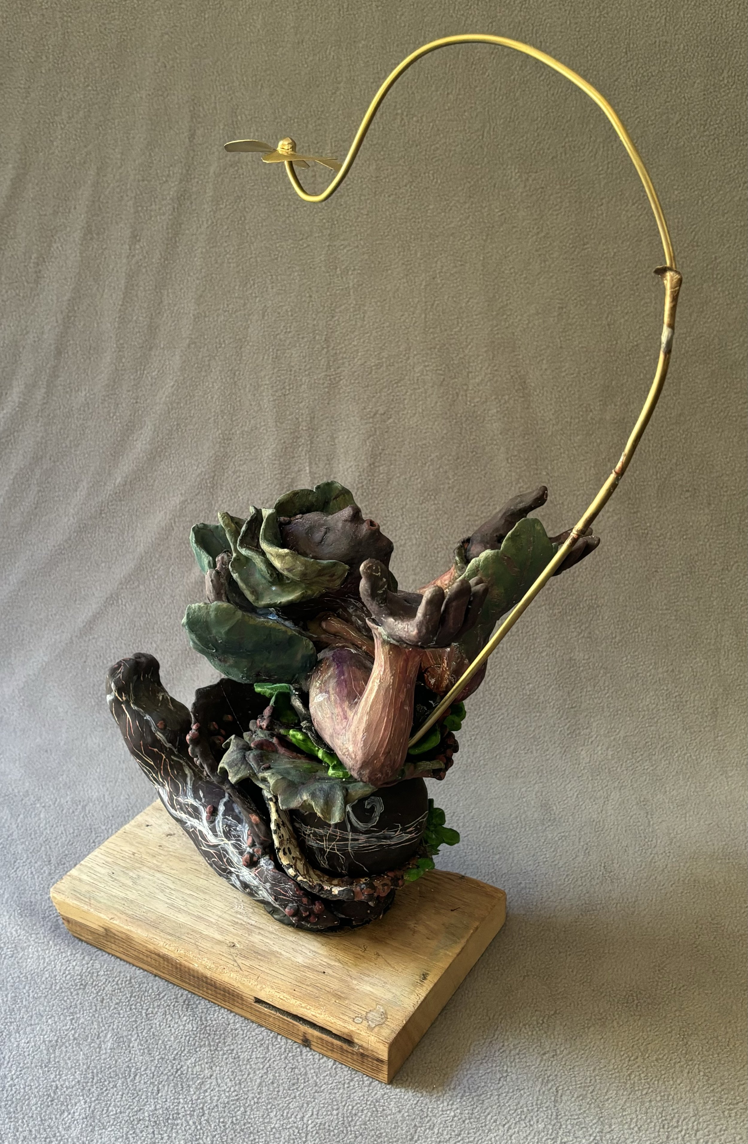 Breath and Breeze, 2024, Ceramic, Brass, Wood and paper 19"Hx10"Wx13"D
Face and hands rise out of a leafy body Kneeling with its feet lifted behind the figure. The body is green, dark gray, black and rust colored. From within the belly area a brass
