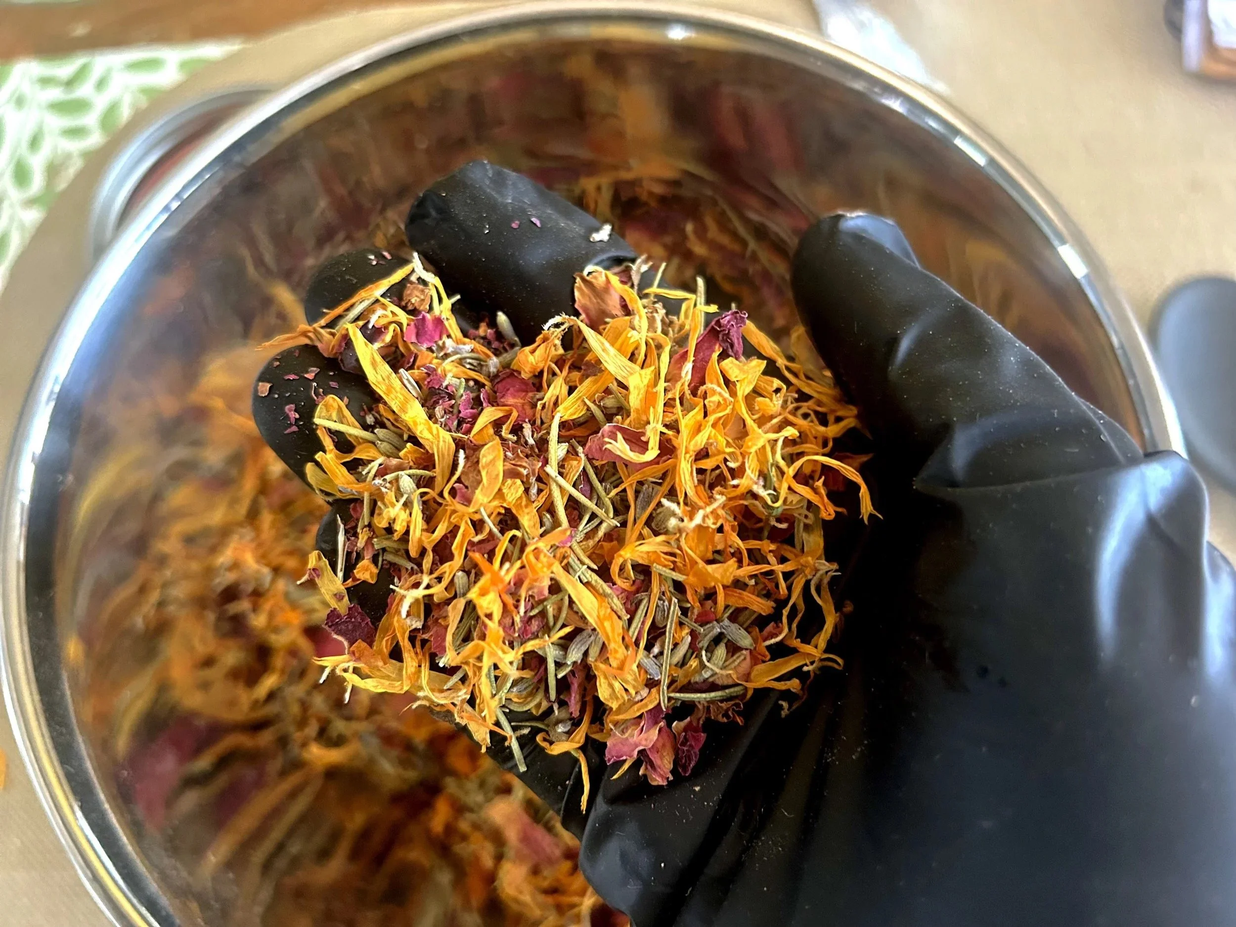 Hand holding a pestle with dried flower petals and herbs in a mortar or bowl, likely for grinding or preparing herbal remedies or tea.
