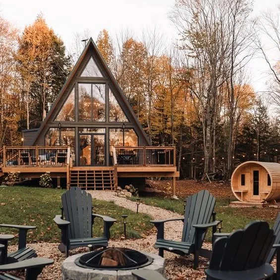 A-Frame glass cabin with a wooden deck, surrounded by trees with autumn leaves, outdoor seating around a fire pit, and a wooden barrel-shaped structure nearby.