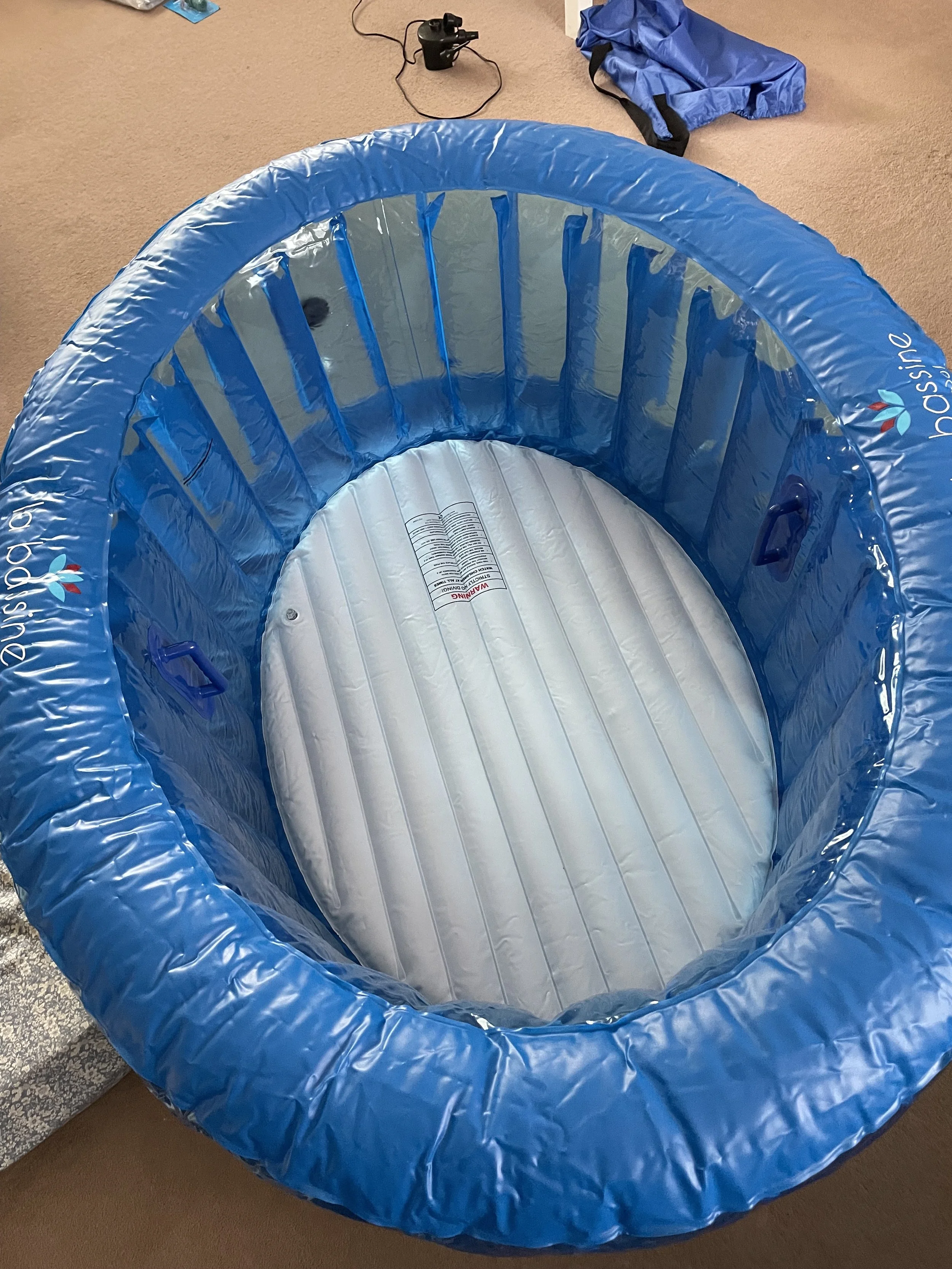 Inflatable baby bassinet with blue outer ring and white interior on beige carpet, with electronic device and folded clothes in background.